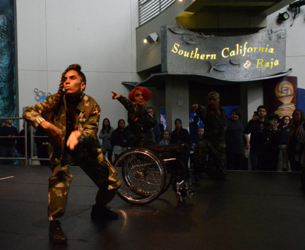 Auti Angel points sternly from her wheelchair, flanked by dancer Ossi, in a crouch in her fatigues, and Ceraldo, pointing towards Ossi