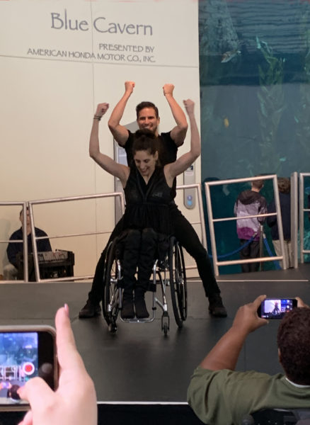 Mia and Marty perform an uptmpo dance with their arms extended upward in unison and his legs extending beyond the back of her wheelchair