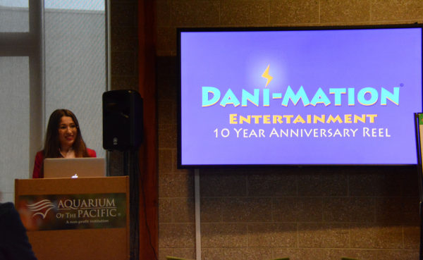Dani smiles from a lectern near a screen with "Dani-Mation Ten Year Anniversary Reel" on it