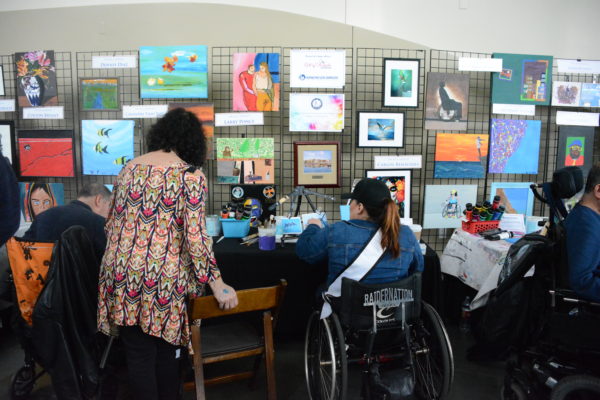Artist with Ms. Wheelchair ribbon on her shirt paints a landscape next to art exhibit