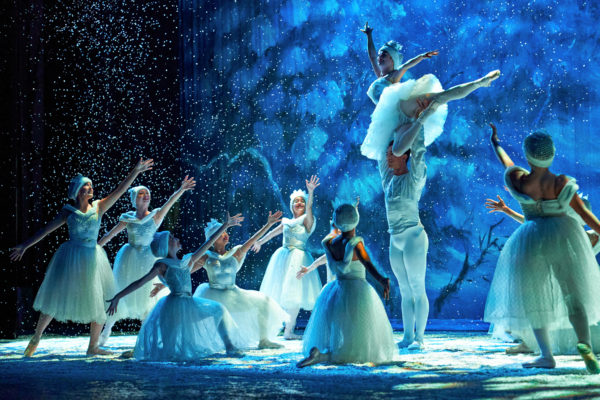 Male ballet dancer lifts ballerina in white tutu and snowy leotards high over his head as other ballerinas dressed as snowflakes surround them and snow falls on a blue-lit stage