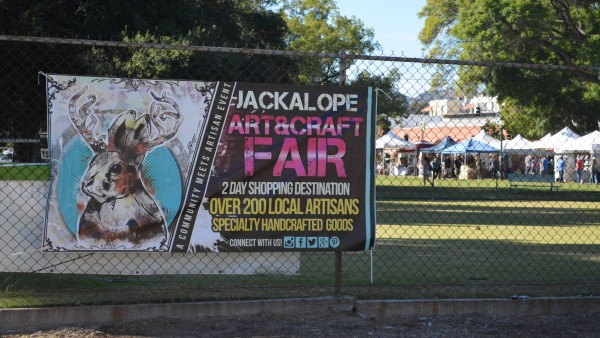 Jackalope Art and Craft Fair sign on chainlink fence at Pasadena's Central Park, with booths set up in the distance.