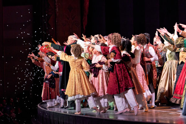 Preteen dancers in party dresses and tights reach towards the edge of the stage as confetti falls