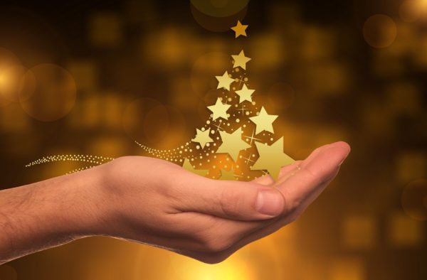Gold stars rise from an extended hand in the shape of a small Christmas tree against a gold blurred background.
