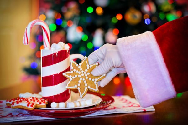 Santa's arm with a white glove, taking a cookie from a plate under a trimmed Christmas tree with a candy-cane-striped mug in the background