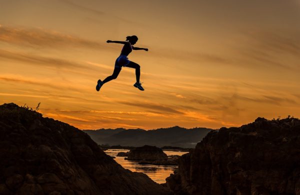 Woman silhouetted against sunset as she leaps from one mountain to another