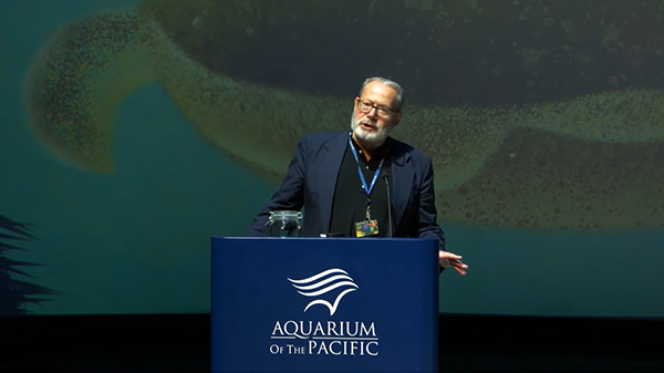 Aquarium of the Pacific President and CEO Dr Peter Karieva speaks from a blue lectern wiht "Aquarium on the Pacific" on it