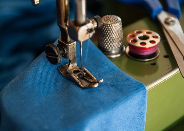 Closep of presser foot on rusted sewing machine, down on a piece of blue cloth, with a metal thimble, bobbin of red thread and pair of scissors nearby
