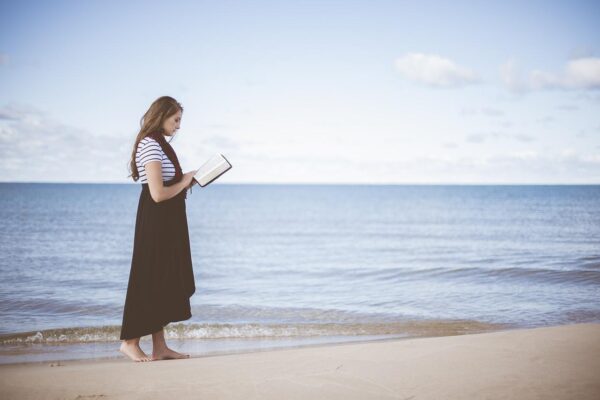 A woman in a long skirt stands barefoot on a beach at the water's edge with a book open as a small wave comes in.