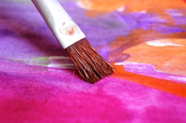 Closeup of paintbrush adding magenta and red paint to an abstract piece with purple and orange areas