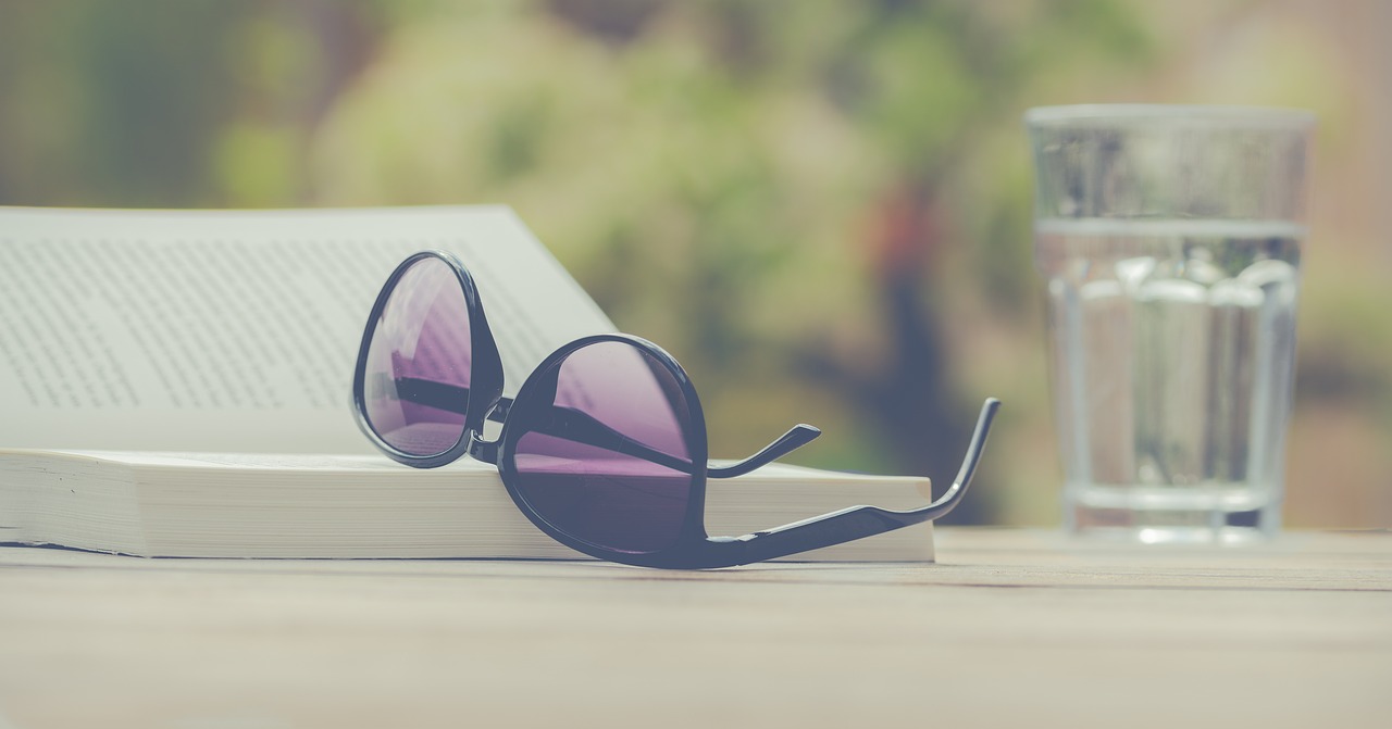 Sunglasses upside down next to a book open on an outdoor table with blurred green trees in the background