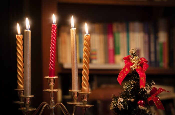 Lighted gold Christmas candles flank a lighted red one in the middle, stand near a small pinecone ornament in front of a shelf full of books.