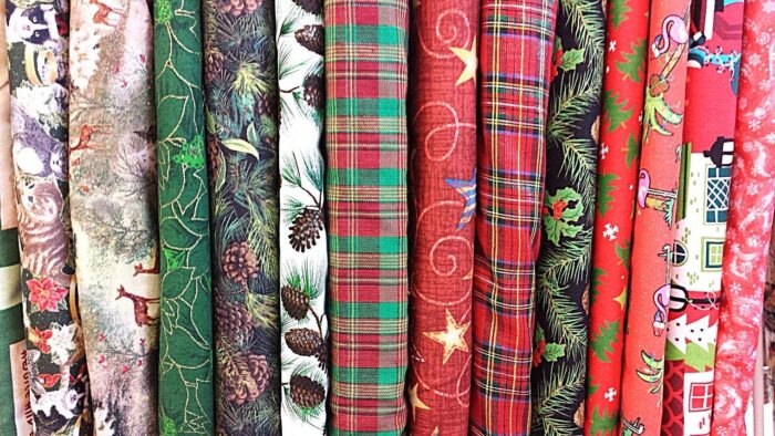 Bolts of Christmas-themed fabric, including red-and-green plaid and a white cotton printed with brown pinecones and green branches, lined up on a shelf.