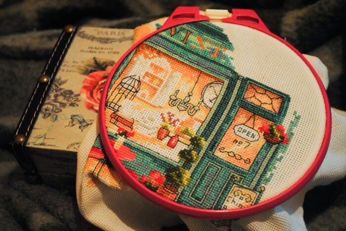 Counted cross stitch picture of a small shop with an "Open" sign in an embroidery hoop