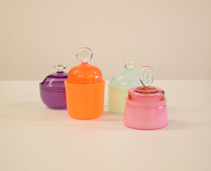 Four lidded glass containers grouped on a counter. Colors are deep purple, orange, light green and rose, all with clear-glass "knob" handles.