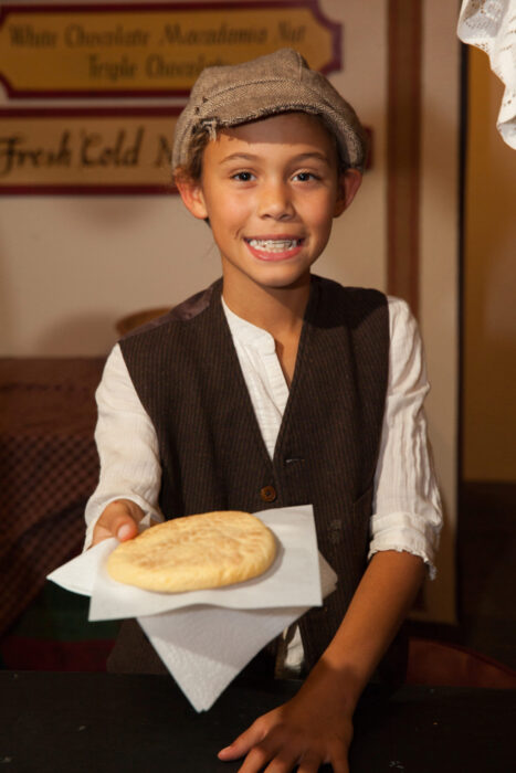 Young boy in newsboy cap and vest with a white shirt offers a large butter cookie.