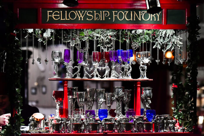 Shop window displays multicolored glassware with heart-shaped clear pedestals and pewter pendants on two shelves under a green sign with a red background and "Fellowship Foundry" in gold lettering.