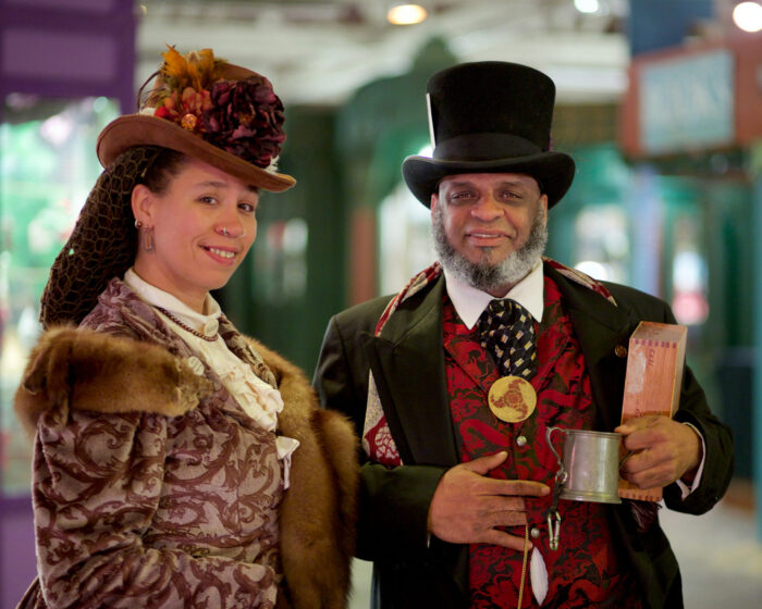 Man in a Victorian hat with a gray jacket and red waistcoat smiles alongside a woman in a Victorian hat and suit
