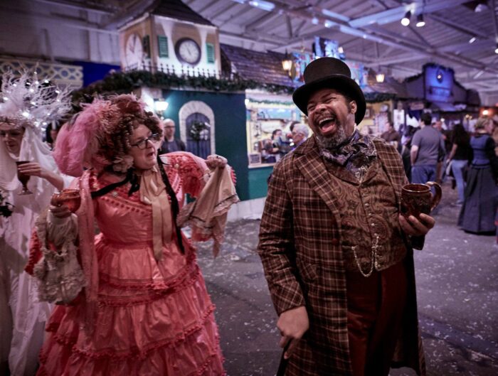 Man in a top hat and Victorian jacket laughs while in conversation with a woman in pink Victorian dress with pink feathers in her hair, on Dickens Fair concourse