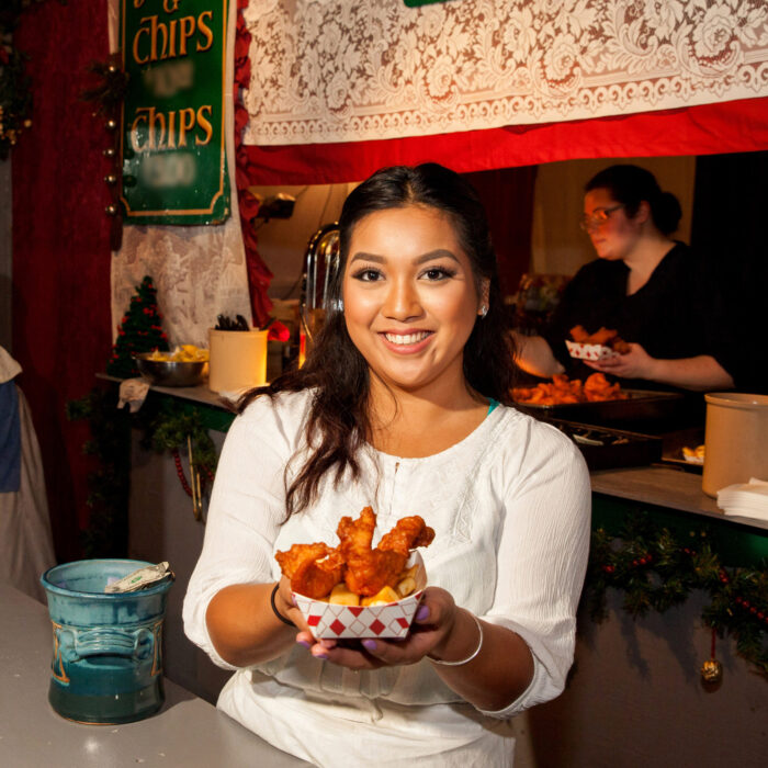 Smiling woman in a white blouse offers a paper container of battered fish and chips next to a green sign reading, "Chips"