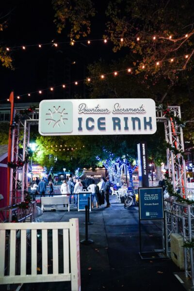 People head for the rink beyond a short white fence as a sign advertises "Downtown Sacramento Ice Rink" under a night sky.