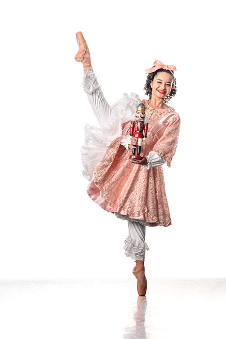 Ballerina in a pink dress, with a pink bow in her hair, en pointe as she holds a nutcracker.