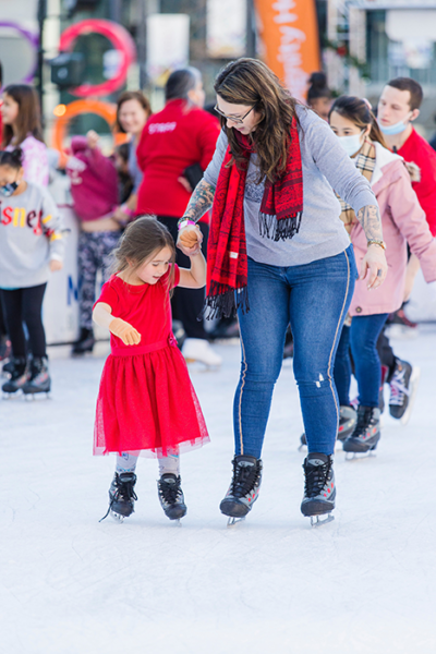 Woman in blue jeans guides a small girl in a red dress as they ice skate together.