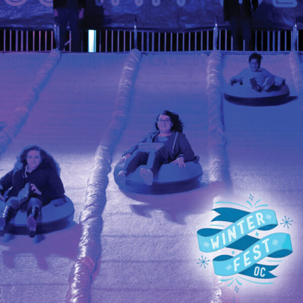 Three smiling participants ice-sled down tracks on a snow-covered "hill" with "Winterfest OC" on a blue banner in the lower right corner of the picture