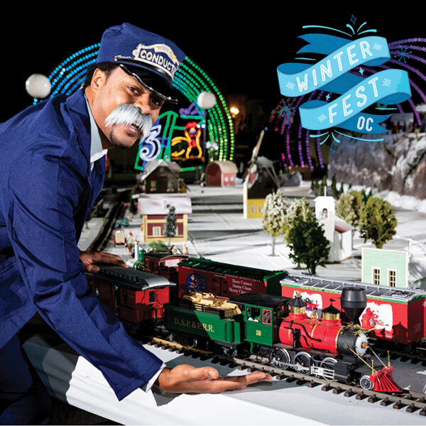 Man in conductor's uniform points to a track of electric trains with a blue banner reading "Winter Fest OC" in the upper right corner of the picture