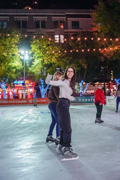 Teenage girl waves from the ice as she and a friend skate under a nighttime sky with overhead lights