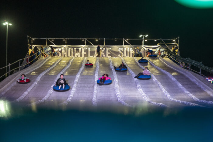 Ice tubers come down slide tracks with "Snowflake Summit" in gold lights behind them