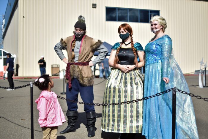 Staffers dressed as Kristoff, Anna and Elsa stand outside the Aerospace Museum of California welcoming a small girl who looks at them in wonder.