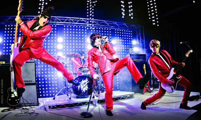 Flashback Heart Attack band, in red 80s-style trousers and jackets, perform dance moves onstage.