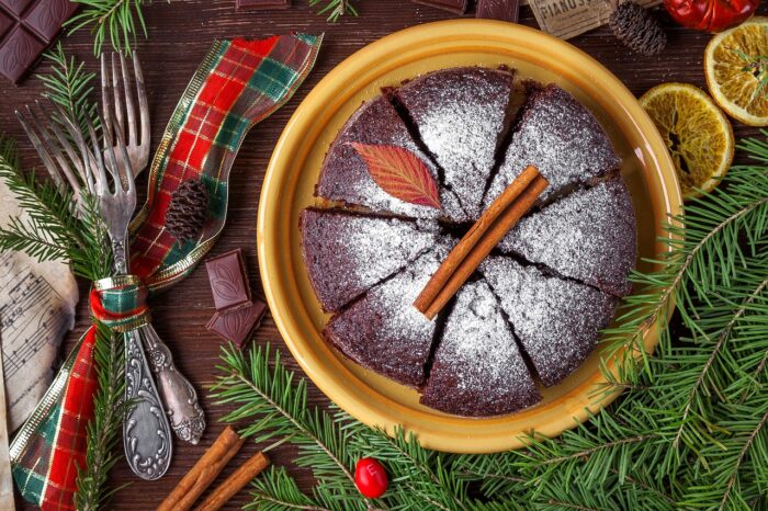 Chocolate Christmas cake with confectioners' sugar topping cut in eight slices, sits on a table near pine branches cinnamon sticks and a red-and-green plaid ribbon.