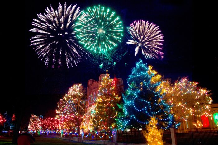 Multicolored fireworks explode starbursts of green, purple and white into the night sky over a forest of lighted Christmas trees in green and gold.