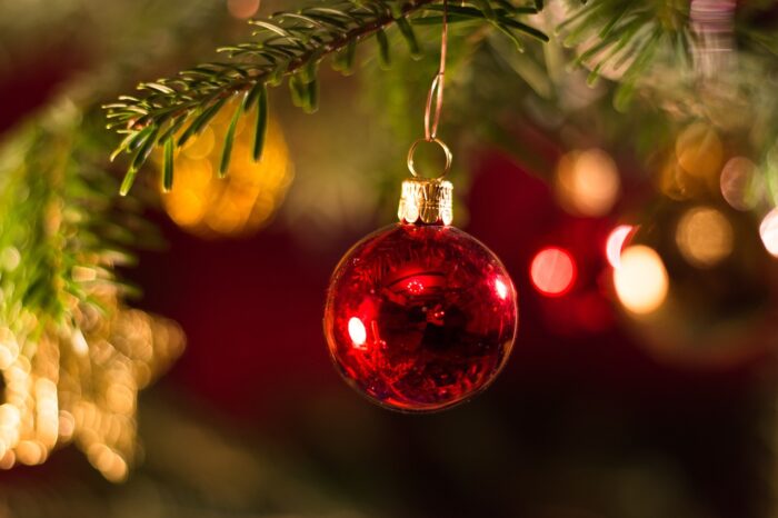 Red Christmas ornament hangs on a pine branch with blurred gold lights in the background.