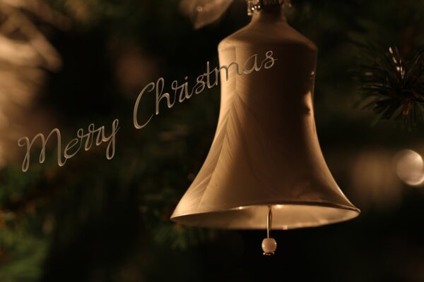 Gold Christmas bell ornament hangs on Christmas tree with "Merry Christmas" lettering over it