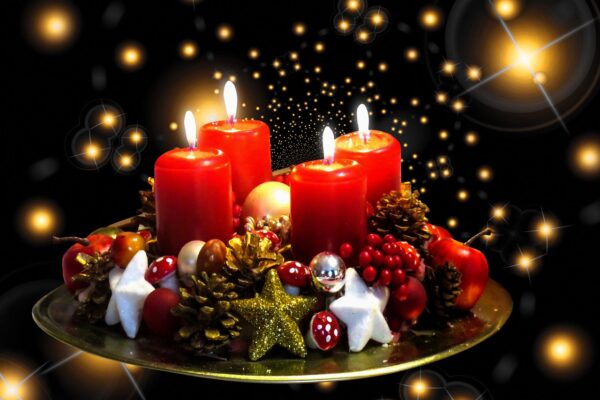 Four lighted red Adventcandles sit on a tray surrounded by pinecones, star ornaments and a red rivvon with pinpoints of light in the background.