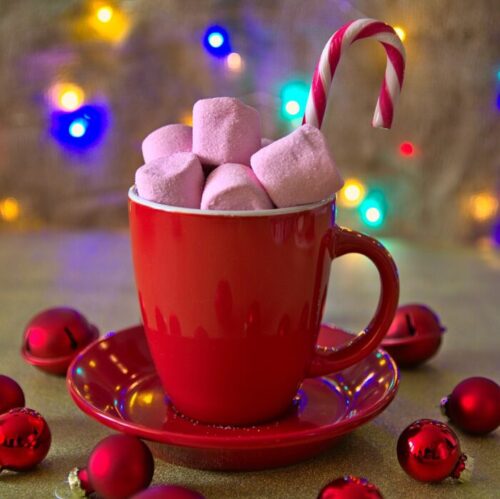 Red mug heaped with marshmallows and a candy cane surrounded by red Christmas ornaments with Christmas lights in the background.