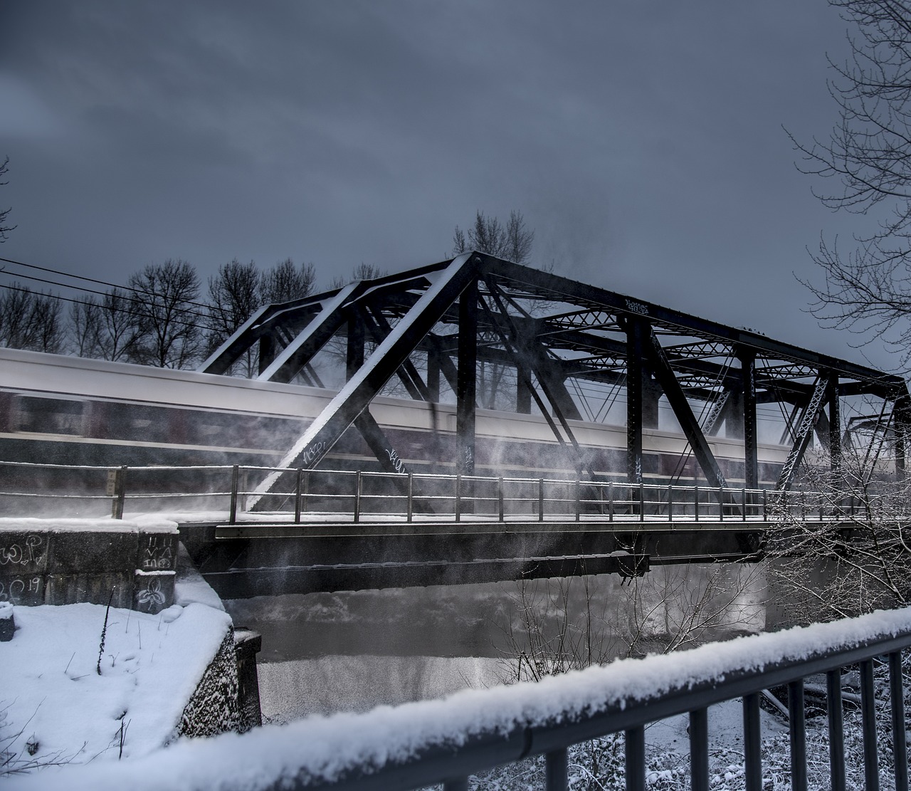 Silver train raises clouds of snow as it goes over railroad trestle in a blur