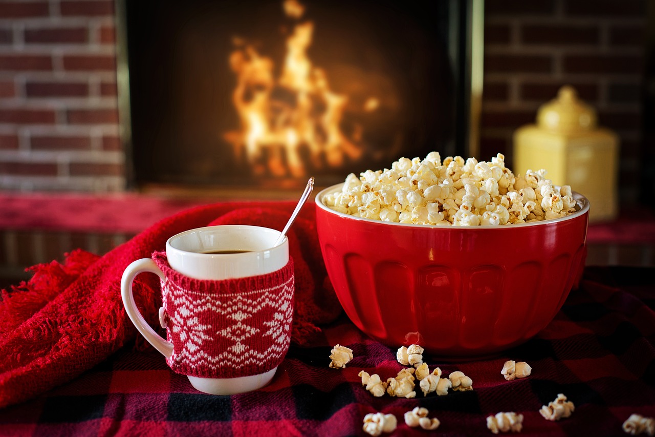 Red bowl of popcorn next to a mug of hot chocolate in a red-and-white cozy near a fireplace with a blazing fire