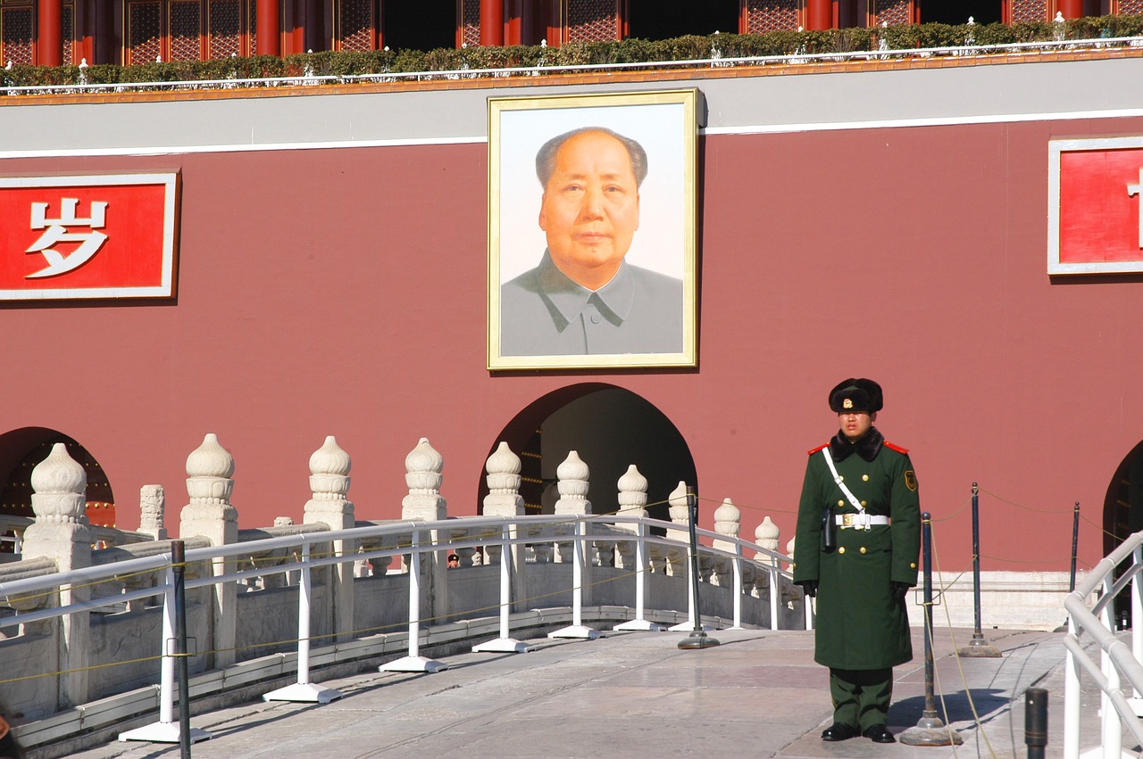 Guard stands on a curved bridge in front of a red-brick building bearing a photo of Mao Zedong.