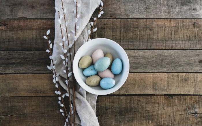 Cup of small candy Easter eggs next to pussy willows against a wooden-board background.