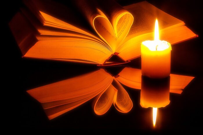 Open book with pages on each side meeting in a heart shape next to a lit candle.