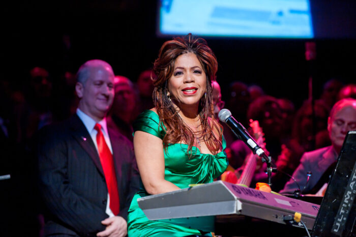 Valerie Simpson, in a green satin gown, sings into a microphone as audience members listen.