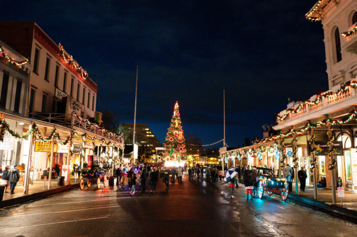Shoppers walk on car-free streets lined by stores festooned with garlands and Christmas lights at night, with the lighted Christmas tree in the background.
