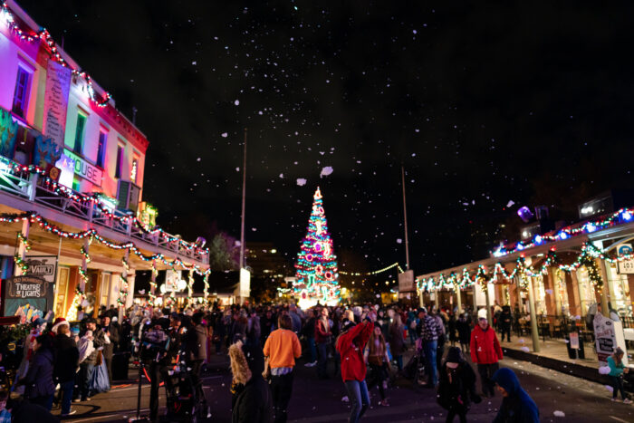 Shoppers at Old Sacramento Waterfront at night, with lighted Christmas tree at the end of a pedestrian area with lighted shops festooned with garlands.