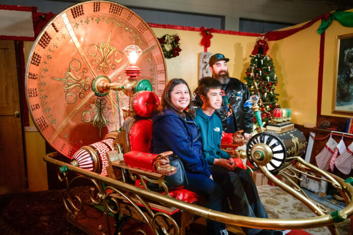 Guest ride in a "Time Machine" with a giant clock face, near a Christmas tree decorated with white candles, as inventor "Dr. Murillo" looks on