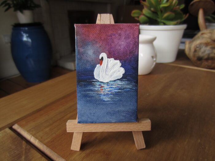 Miniature painting of a swan sits on a small easel on a desk.