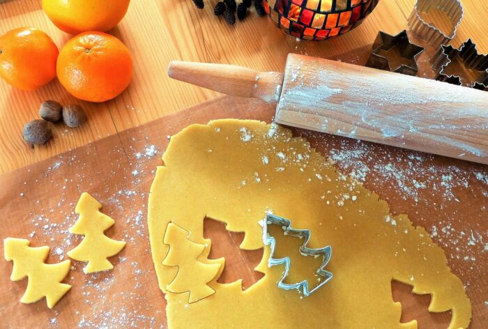 Floured rolling pin sits on rolled-out cookie dough with a tree-shaped cookie cutter and oranges and a candle in the background.